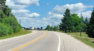 Two-lane road curving ahead, green trees lining both sides, blue sky with fluffy clouds